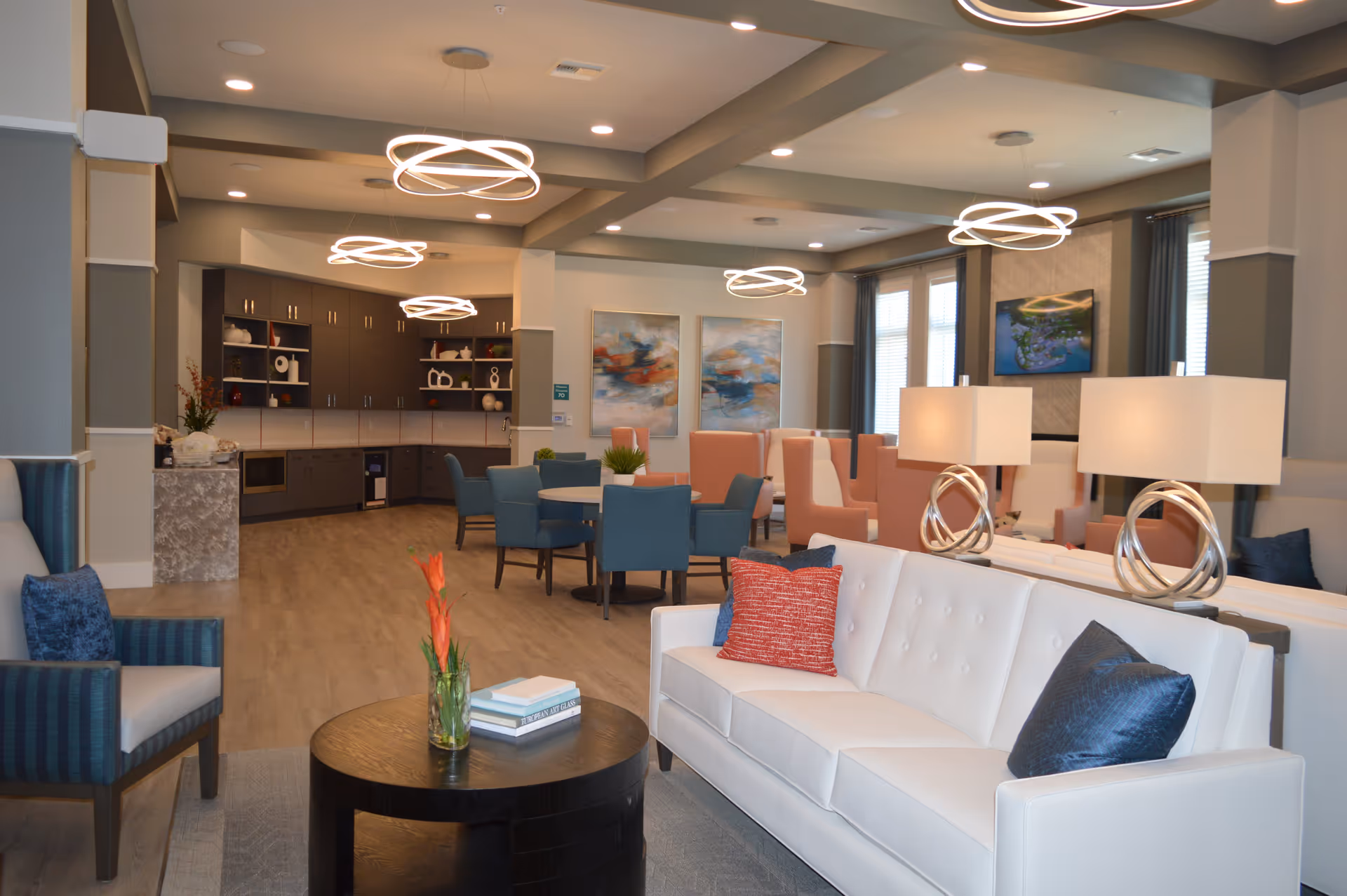 A modern senior living common area with a white sofa adorned with red and blue pillows, a round wooden coffee table with a vase of orange flowers and books, blue and pink chairs around tables, contemporary circular ceiling lights, and a kitchenette with dark cabinets in the background.