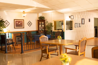 Interior view of a common area in a senior living facility with wooden tables and chairs, decorative plants, framed artwork on the walls, and a small fireplace in the background.
