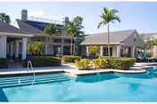Outdoor swimming pool area with clear blue water, surrounded by a patio and landscaped bushes. Two single-story buildings with gray roofs and palm trees are visible in the background under a clear sky.