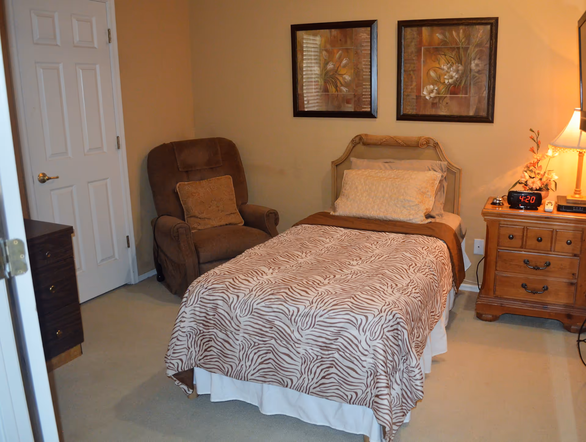 A cozy bedroom with a single bed covered in a zebra-striped blanket and multiple pillows. Next to the bed is a wooden nightstand with a lamp, a digital clock showing 4:20, a small flower arrangement, and a bell. A comfortable brown armchair with a cushion is positioned in the corner near a closed white door. Two framed floral paintings hang on the beige wall above the bed.