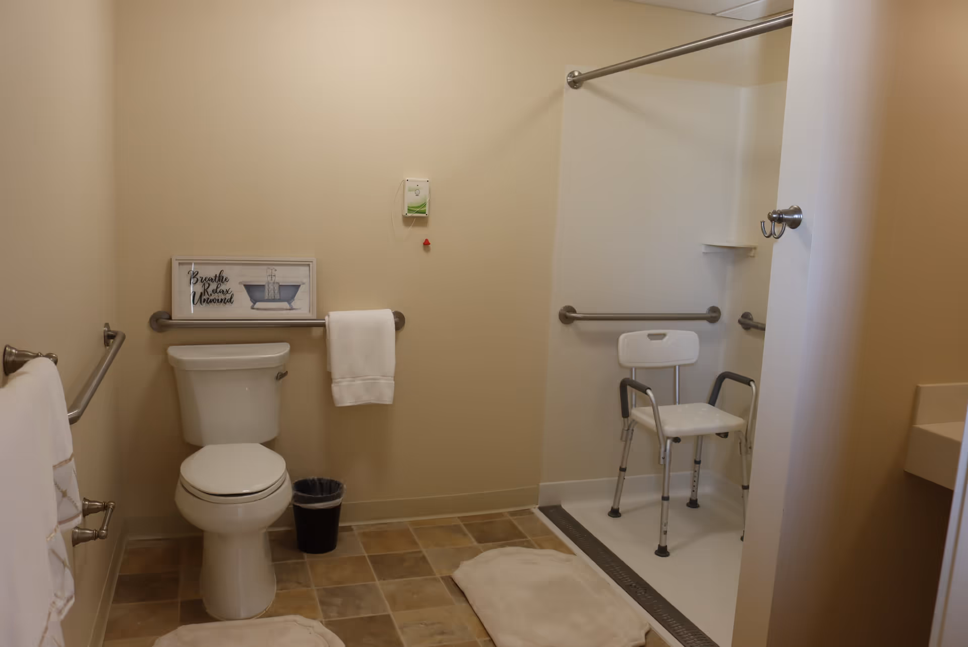 A clean and accessible bathroom with beige walls and tiled floor. It features a toilet with a towel hanging on a grab bar above it, a small trash bin, and a walk-in shower area with a white shower chair and multiple grab bars. There are two beige bath mats on the floor and a framed sign on the wall that reads 'Breathe Relax Unwind'.