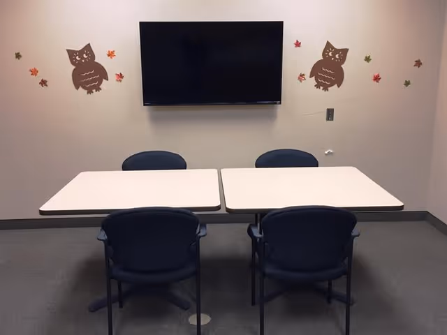 A small meeting or activity room with two white tables pushed together and four blue chairs around them. A flat-screen TV is mounted on the beige wall above the tables. The wall is decorated with brown owl decals and colorful autumn leaves.
