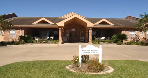 Front exterior view of a single-story brick building with a peaked roof and a covered entrance. There are two chairs on the porch, shrubs and plants along the front, and a sign in the foreground that reads 'Homestead Residence Independent Living'.