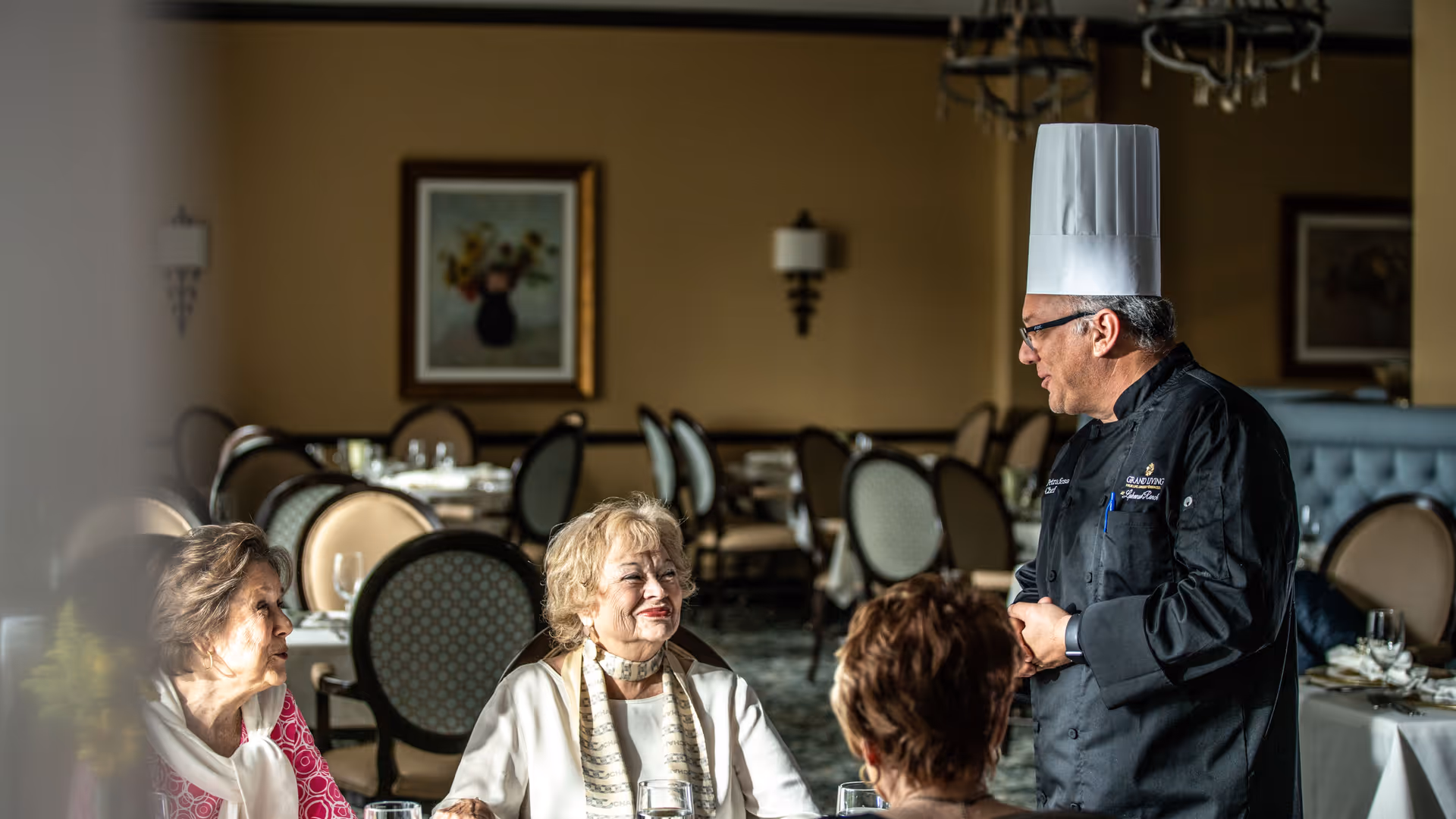 A chef wearing a white chef hat and black uniform is standing and talking to three elderly women seated at a dining table in a restaurant setting with elegant chairs and tables in the background.