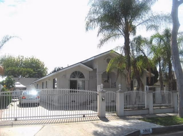 Exterior view of a single-story residential building with a gated driveway and several palm trees in front. The building has a light-colored facade and a sloped roof.