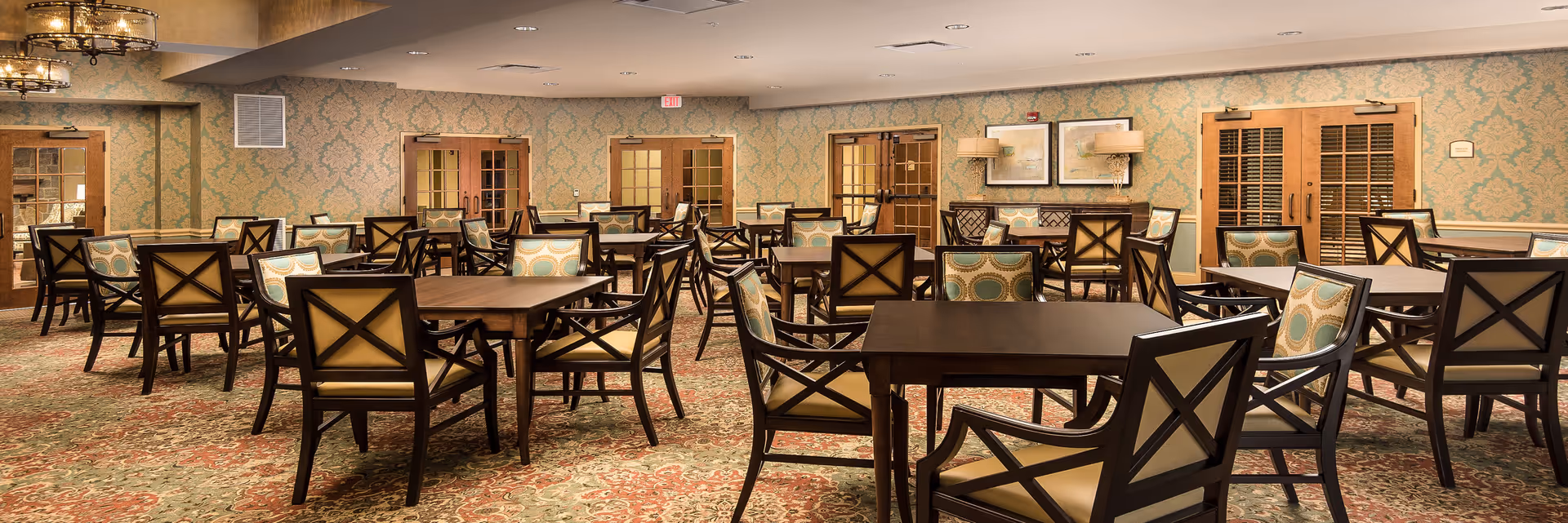A spacious dining room with multiple dark wood tables and chairs featuring patterned cushions. The room has ornate wallpaper with a damask pattern, a patterned carpet, and several wooden double doors with glass panels. The lighting includes ceiling fixtures and wall lamps, and there are framed artworks on the walls.