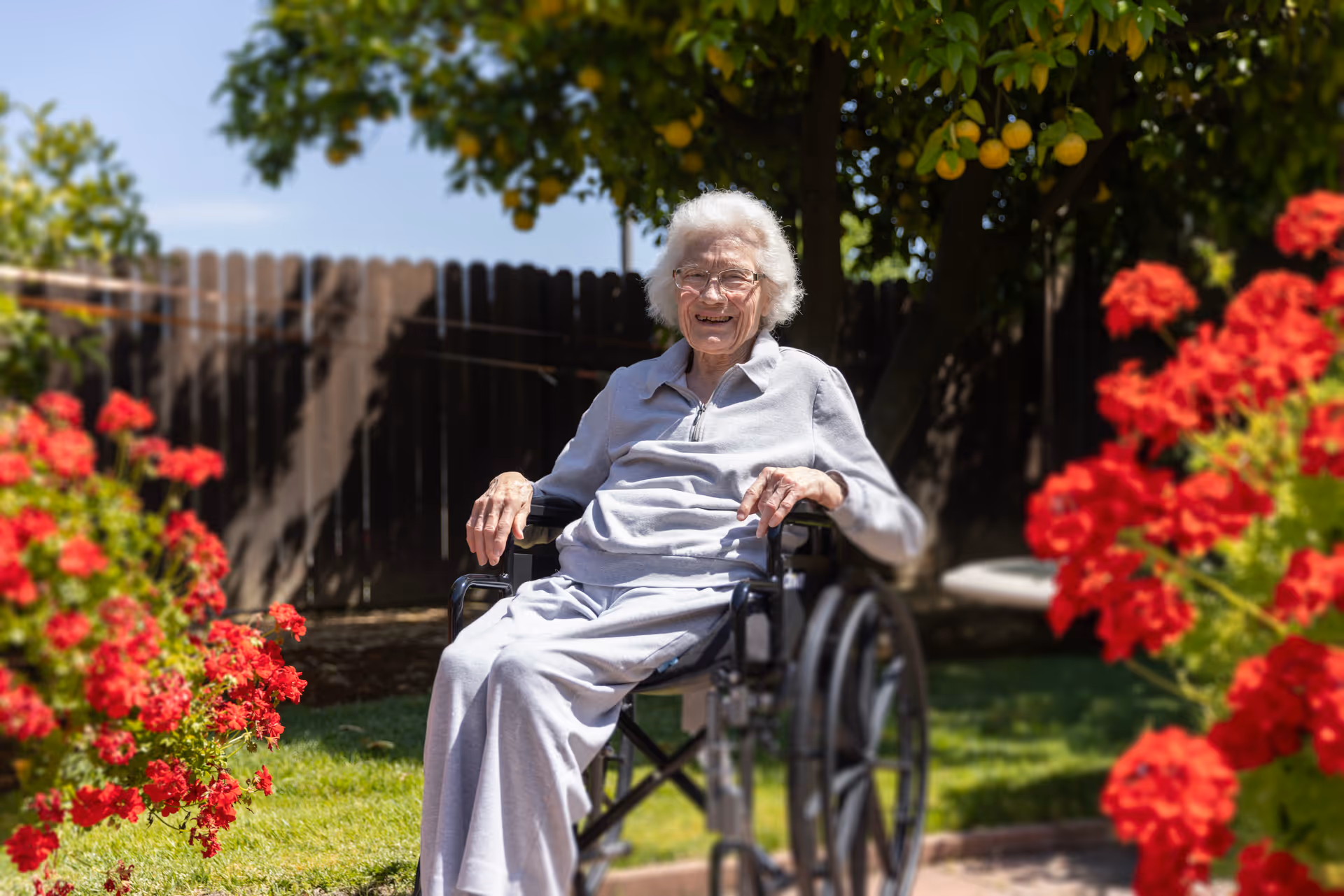 An elderly woman in a wheelchair smiling outdoors in a garden with red flowers and a lemon tree in the background, under a clear blue sky.