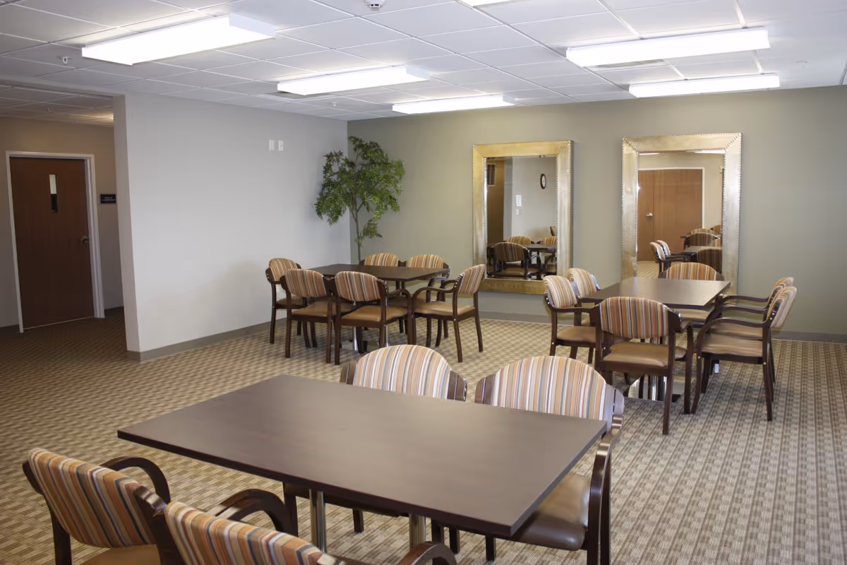 A common area with multiple dark wood tables and striped cushioned chairs arranged around them. The room has a patterned carpet, two large decorative mirrors on one wall, a potted artificial tree in the corner, and fluorescent ceiling lights. There is a wooden door and a hallway visible in the background.
