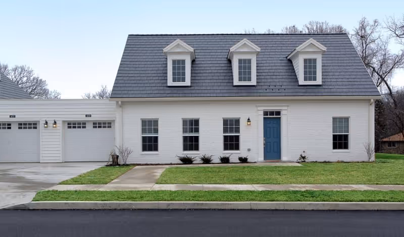 Front exterior view of a white two-story house with a dark gray roof, three dormer windows, a blue front door, and an attached garage with two doors. The house is surrounded by a green lawn and a concrete driveway and sidewalk.