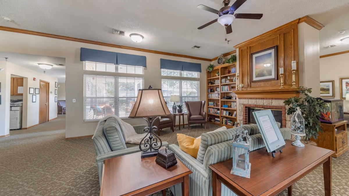 A cozy living room area in a senior living facility featuring a striped sofa with yellow and gray cushions, a wooden side table with a decorative lamp, a wooden coffee table with framed information and decorative items, two brown leather armchairs near large windows with blue valances, a built-in wooden bookshelf filled with books, and a brick fireplace with a wooden mantle and framed artwork above it. The room has beige walls, carpeted floors, and a ceiling fan with light.