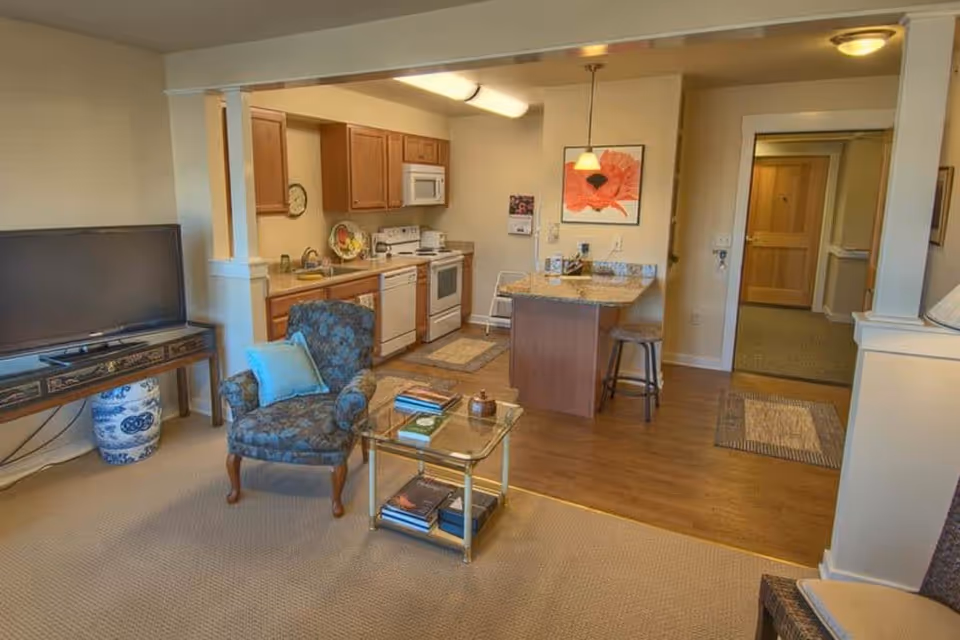 Interior view of a senior living facility apartment at Chandler's Square showing a combined living room and kitchen area. The living room has a patterned armchair with a blue pillow, a glass coffee table with books, and a TV on a wooden stand. The kitchen features wooden cabinets, a white stove, microwave, dishwasher, and a granite countertop island with a bar stool. The flooring transitions from carpet in the living area to wood in the kitchen. A framed flower artwork hangs on the wall near the kitchen island.