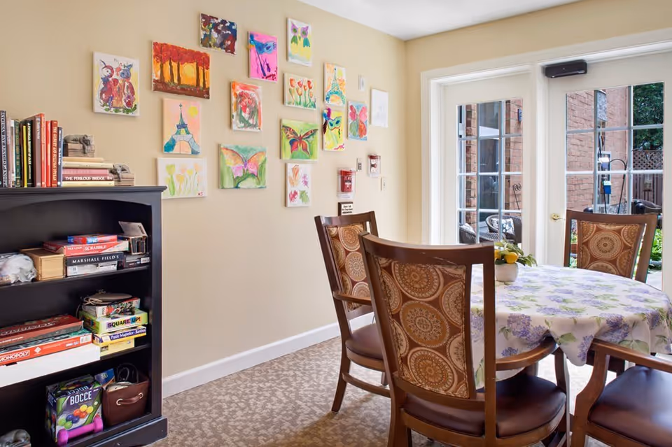 A cozy dining area with a round table covered with a floral tablecloth and four wooden chairs with patterned upholstery. On the left side, there is a black bookshelf filled with books and board games. The wall above the bookshelf is decorated with colorful paintings, mostly of butterflies and flowers. French doors on the right open to an outdoor patio area with brick walls and garden furniture.