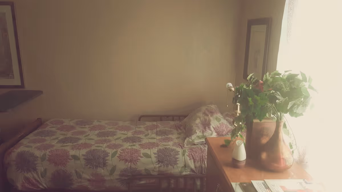 A small bedroom with a single bed covered in a floral patterned bedspread in shades of purple and green. Next to the bed is a wooden dresser with a large vase filled with green leafy plants, a small white object, and some papers or magazines. The room has soft natural light coming through a window with sheer curtains on the right side.
