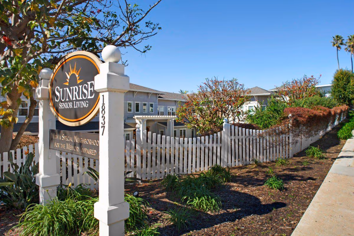 Exterior view of Sunrise Senior Living facility with a white picket fence, landscaped garden, and clear blue sky. The sign reads 'Sunrise Senior Living' with additional text about a special neighborhood for the memory impaired.