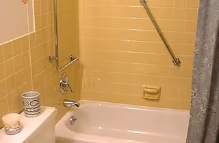 Bathroom with yellow tiled walls, a white bathtub with a silver faucet and showerhead, a grab bar on the wall, a soap holder, a white toilet with a candle and a decorative container on top, and a gray shower curtain partially visible on the right.