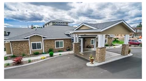 Exterior view of Fieldstone Memory Care Yakima showing a single-story building with a covered entrance supported by stone pillars, surrounded by a paved driveway and some landscaping with shrubs and plants under a partly cloudy sky.