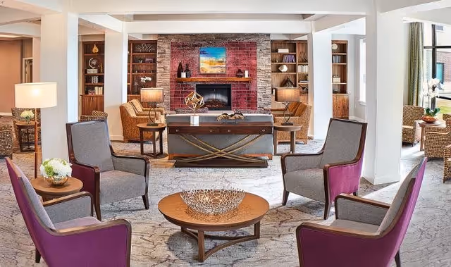 A bright and spacious living room area in an assisted living facility featuring a seating arrangement with four purple and gray armchairs around a round wooden coffee table with a decorative bowl. In the background, there is a brick fireplace with a colorful painting above it, flanked by built-in wooden shelves with decorative items and lamps on side tables. Large windows on the right side let in natural light.