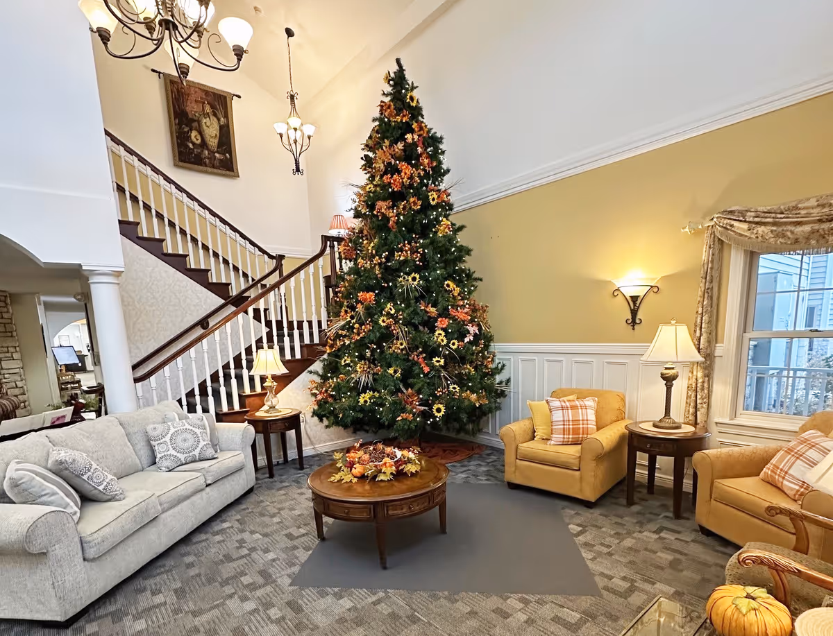 A cozy living room area decorated for the holidays with a tall Christmas tree adorned with orange and yellow ornaments. The room features a beige sofa with patterned pillows, two yellow armchairs with plaid pillows, a round wooden coffee table with a fall-themed centerpiece, and a staircase with wooden handrails and white balusters. Warm lighting is provided by table lamps and wall sconces, and a window with floral curtains lets in natural light.