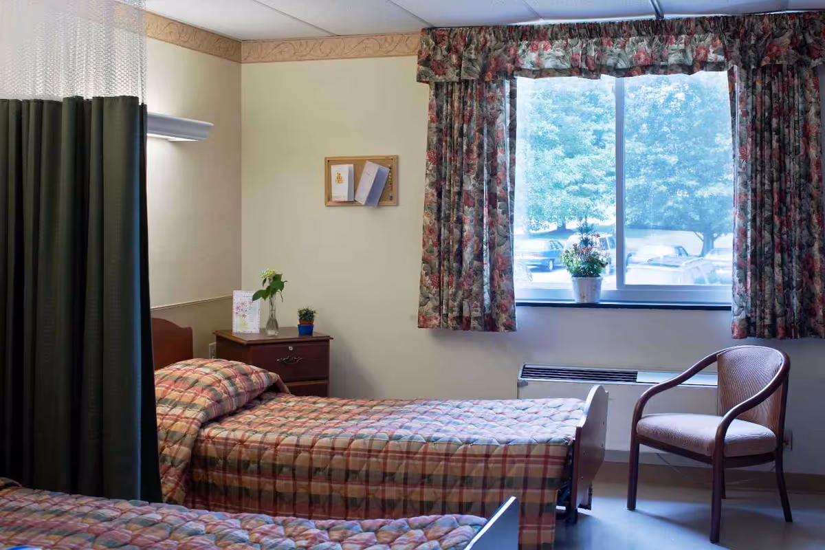 A senior living facility bedroom with two single beds covered in plaid bedspreads, a wooden nightstand with a vase and a small plant, a window with floral curtains showing trees and parked cars outside, and a wooden chair near the window.