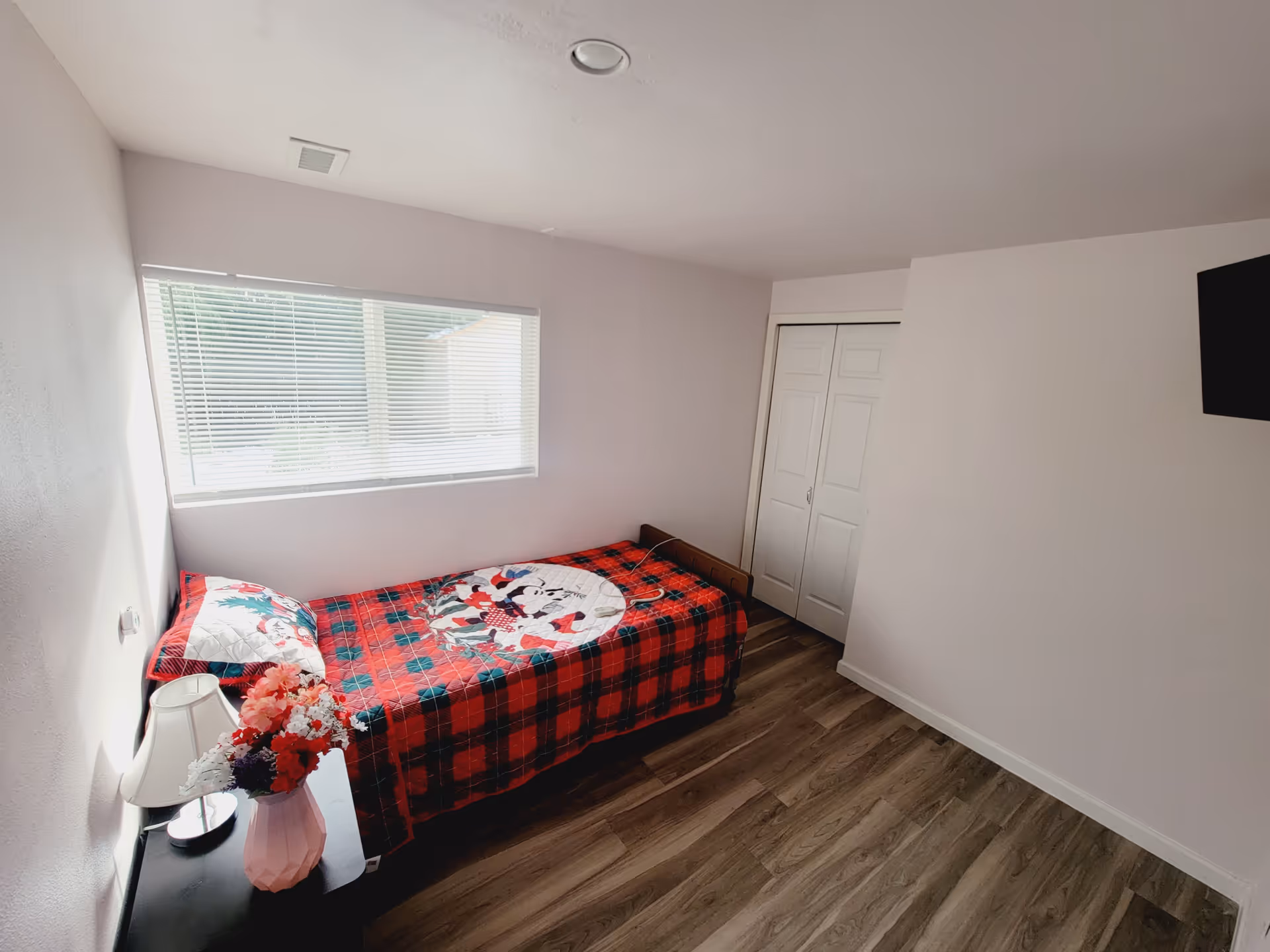 A small bedroom with a single bed covered in a red and black plaid blanket featuring a Mickey Mouse design. Next to the bed is a black nightstand with a white lamp and a pink vase holding a bouquet of colorful flowers. The room has light-colored walls, a window with blinds, a closet with white double doors, and wood-patterned flooring.