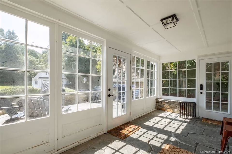 A bright sunroom with large white-framed windows and glass doors allowing natural light to fill the space. The floor is made of stone tiles, and there are a few small rugs on the floor. Outside, greenery and a small white shed are visible.