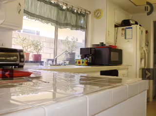 A bright kitchen with white tiled countertops and cabinets. There is a window with green curtains above the sink, showing potted plants outside. On the counter, there is a toaster oven, a coffee maker, a microwave, and a refrigerator with magnets and notes attached.