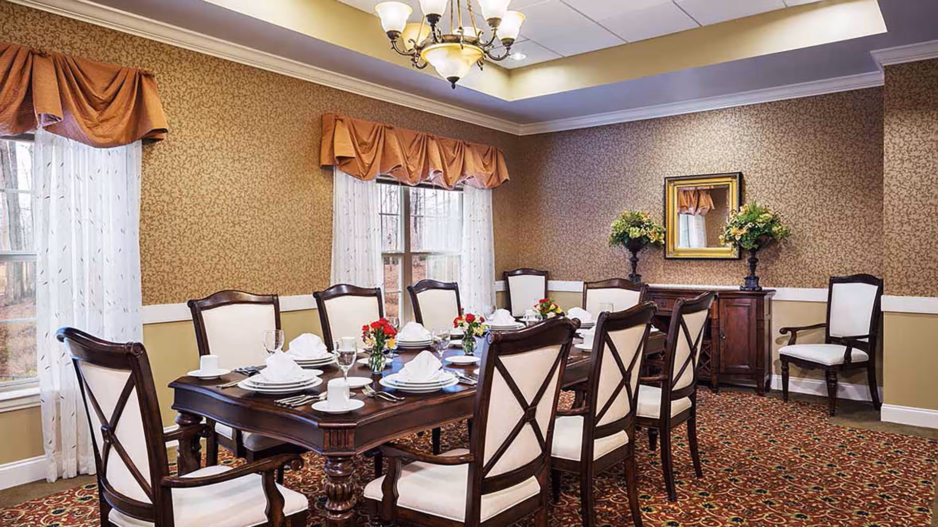 A formal dining room with a large wooden table set for eight people. The table is adorned with white plates, napkins, cups, glasses, and small floral centerpieces. The room has patterned wallpaper, two windows with sheer white curtains and brown valances, a chandelier hanging from the ceiling, and a sideboard with a mirror and flower arrangements.