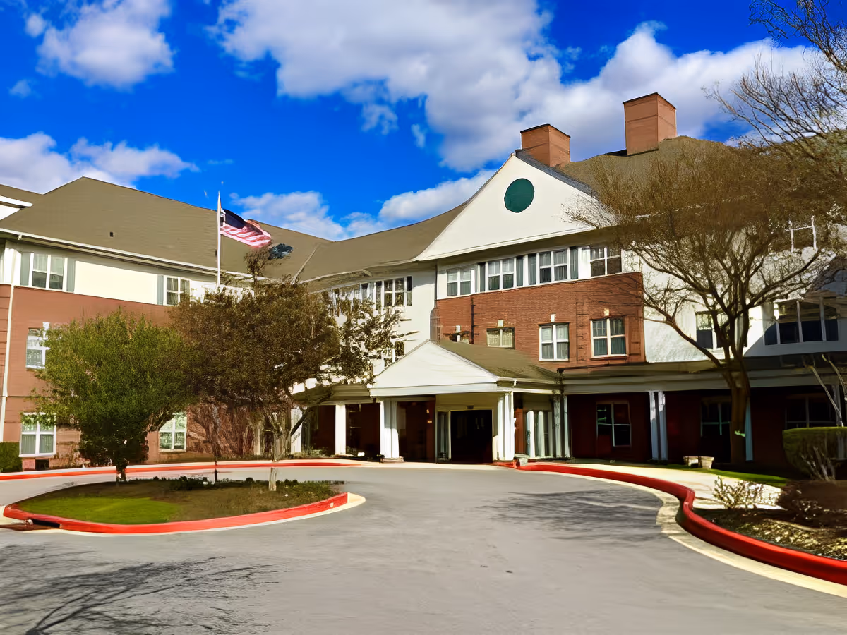 Exterior view of Charter Senior Living of Towson building with a circular driveway, trees, and an American flag flying on a flagpole under a partly cloudy blue sky.