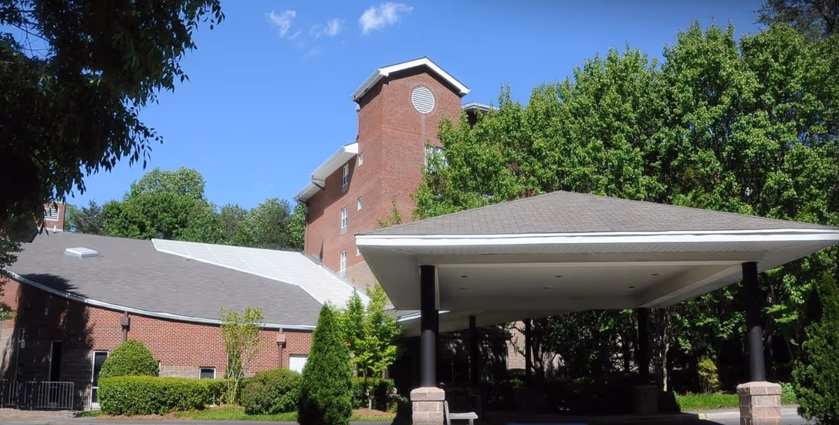 Exterior view of a multi-story brick building with a covered entrance area supported by black columns. The building is surrounded by green trees and bushes under a clear blue sky.