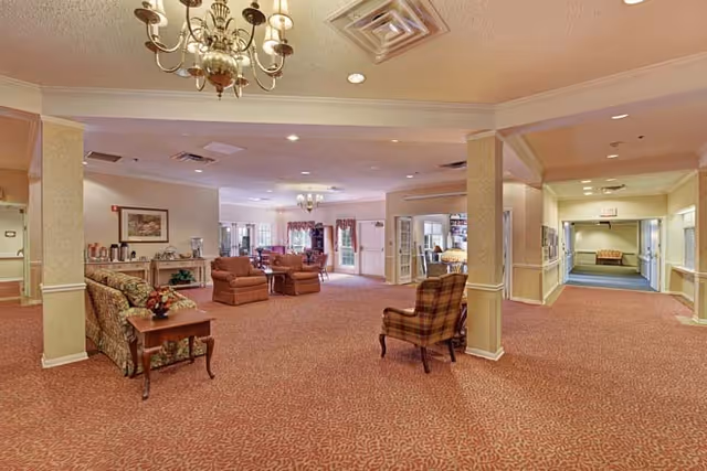 Spacious senior living facility common area with patterned carpet, multiple seating arrangements including sofas and armchairs, chandeliers, and a hallway leading to other rooms.