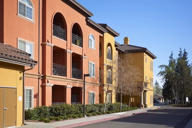 Exterior view of a multi-story senior living facility building with balconies, arched windows, and a tiled roof. The building is painted in warm colors including orange and yellow. There are trees and shrubs along the sidewalk and a clear blue sky above.