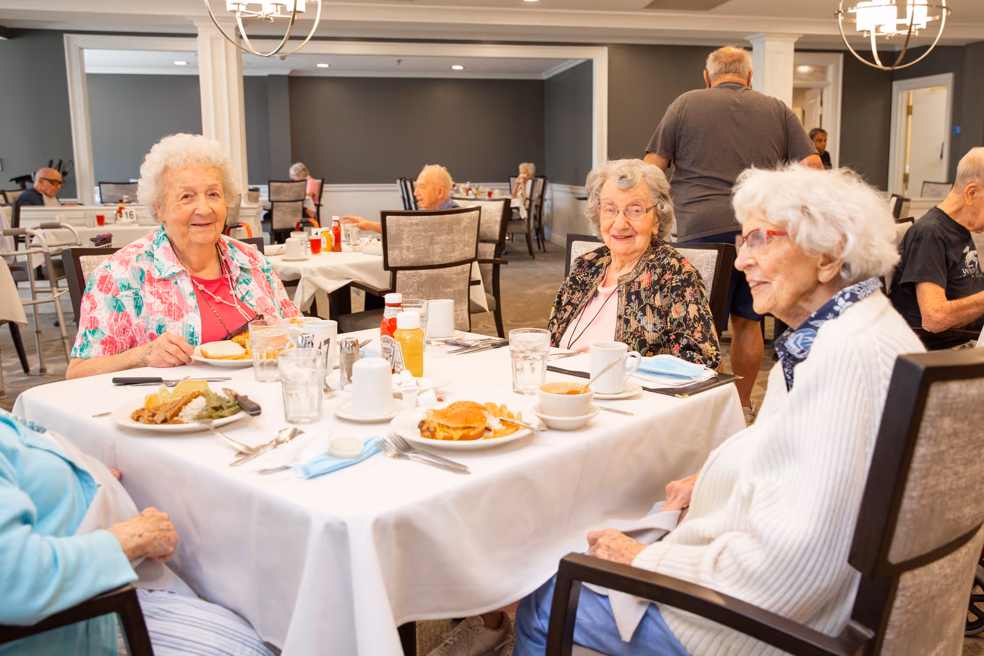 A group of elderly women sitting around a table in a dining room, enjoying a meal together. The table is set with plates of food, glasses of water, coffee cups, and condiments. Other seniors are visible dining in the background in a well-lit, spacious room with neutral-colored walls and modern lighting fixtures.