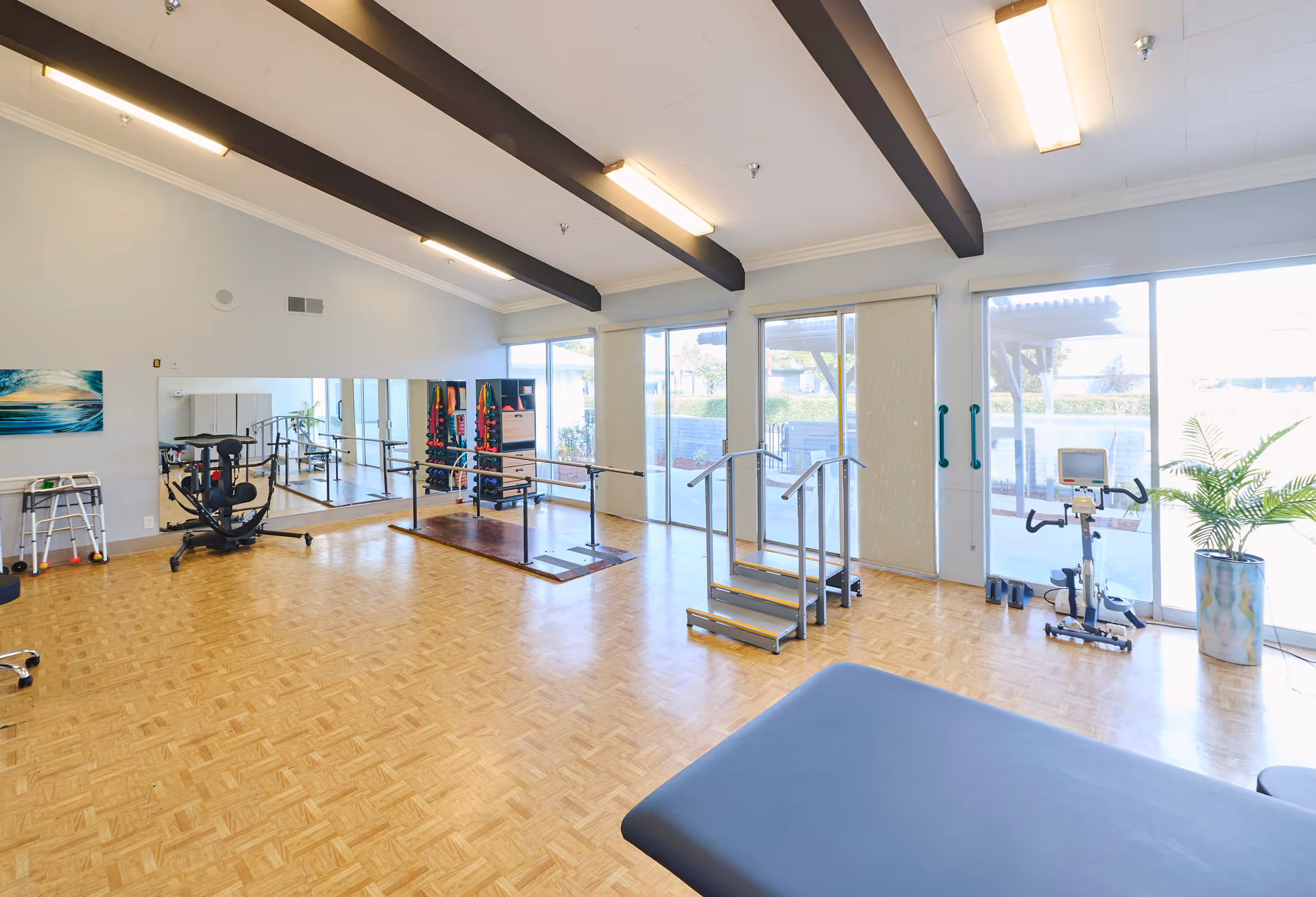 Spacious therapy and exercise room with wooden floors, parallel bars, exercise equipment, and large windows letting in natural light.