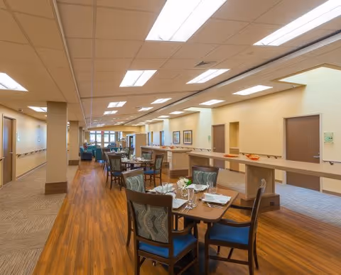 Well-lit senior living dining area with multiple set tables and chairs, wood floors, and a serving counter along a central corridor.