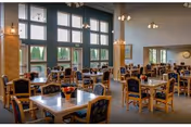 Spacious dining room with multiple wooden tables and chairs arranged neatly. Large windows with black frames allow natural light to fill the room. The walls are light-colored, and there are several light fixtures hanging from the ceiling. A small flower arrangement is placed on each table.