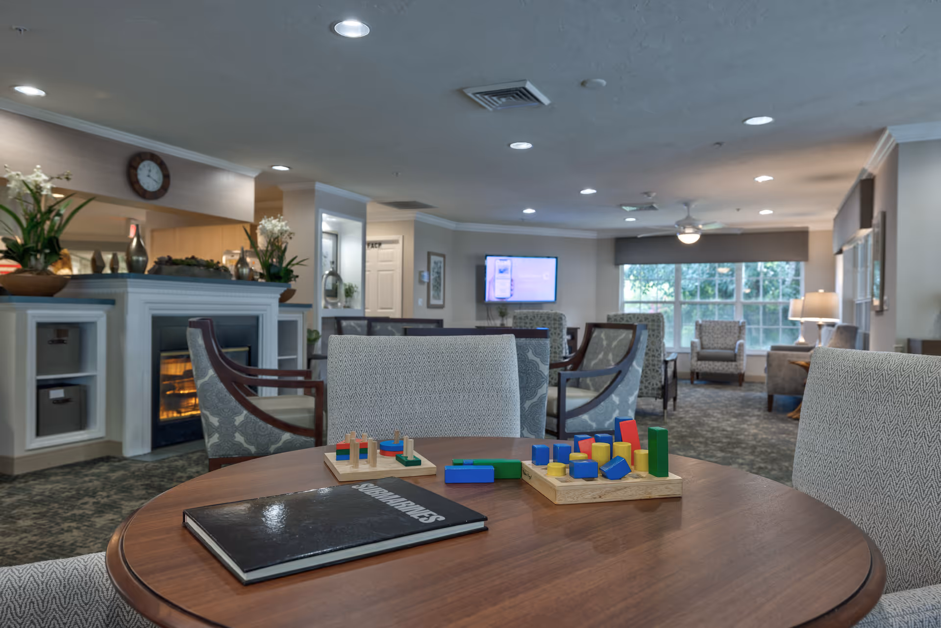 A cozy senior living common area with a round wooden table in the foreground featuring colorful wooden block toys and a book titled 'Senior Games'. The room has several upholstered chairs, a fireplace with decorative plants on the mantle, a wall-mounted TV, and large windows letting in natural light.