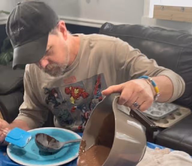 A man wearing a black cap and a Marvel superhero shirt is sitting on a black leather couch, pouring chocolate batter from a mixing bowl into a chocolate mold on a blue plate. A muffin tray is visible in the background.