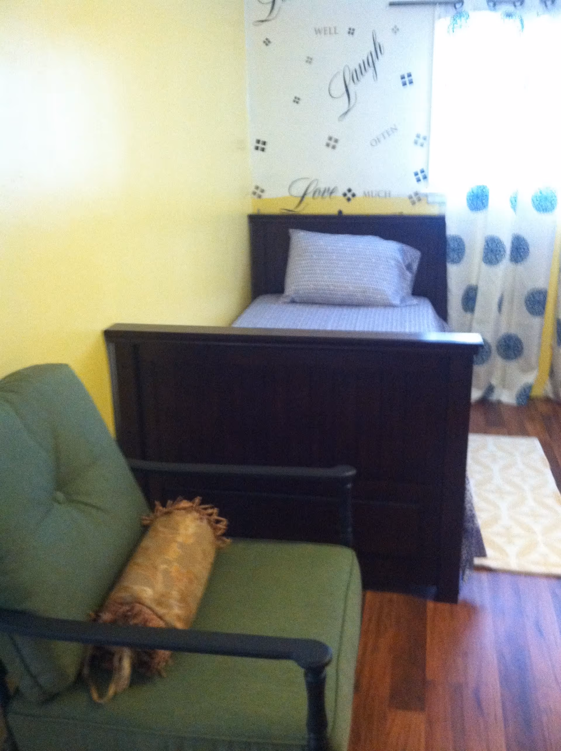 A small bedroom with a single dark wooden bed against a yellow wall. The bed has a pillow and a light-colored bedspread. Next to the bed is a green cushioned chair with a decorative cylindrical pillow. The floor is wooden, and there is a window with white curtains featuring blue circular patterns. Above the bed, there is a wall decal with the words 'Live Well, Laugh Often, Love Much.'