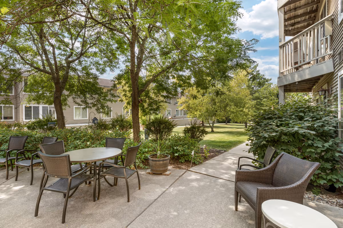 Outdoor patio area at Layton Terrace Senior Living with a round table and six chairs, a wicker loveseat, and a small round side table. The patio is surrounded by green trees, bushes, and a well-maintained lawn with a sidewalk leading through the garden. Residential buildings are visible in the background under a partly cloudy sky.