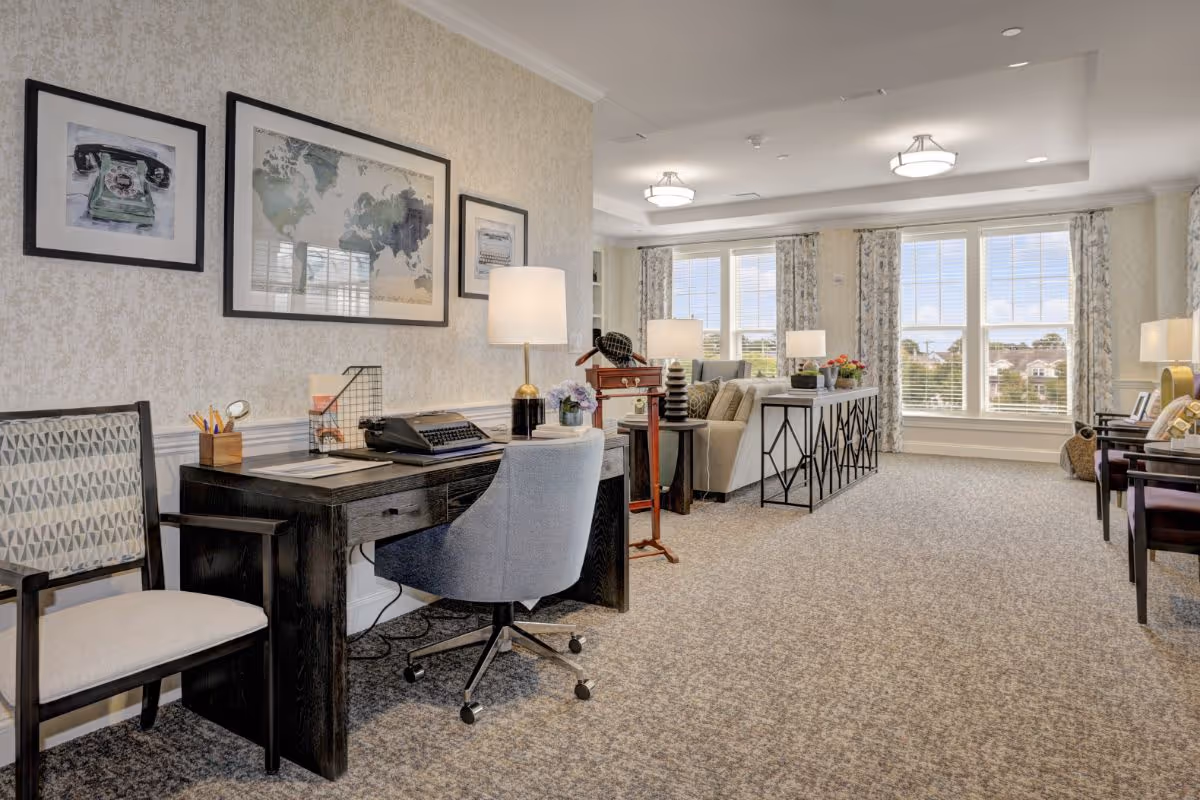 Well-lit senior living common area with a desk and chair in the foreground and sofas and large windows in the background.
