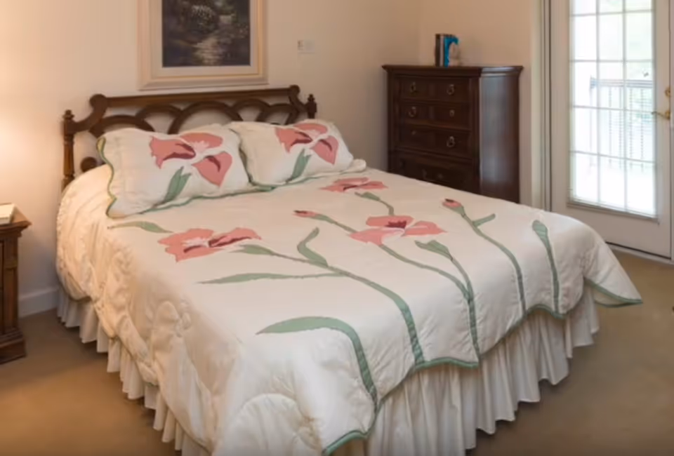 Queen bed with a white quilted bedspread featuring pink flowers, wooden headboard, dresser and a glass patio door in a bedroom.