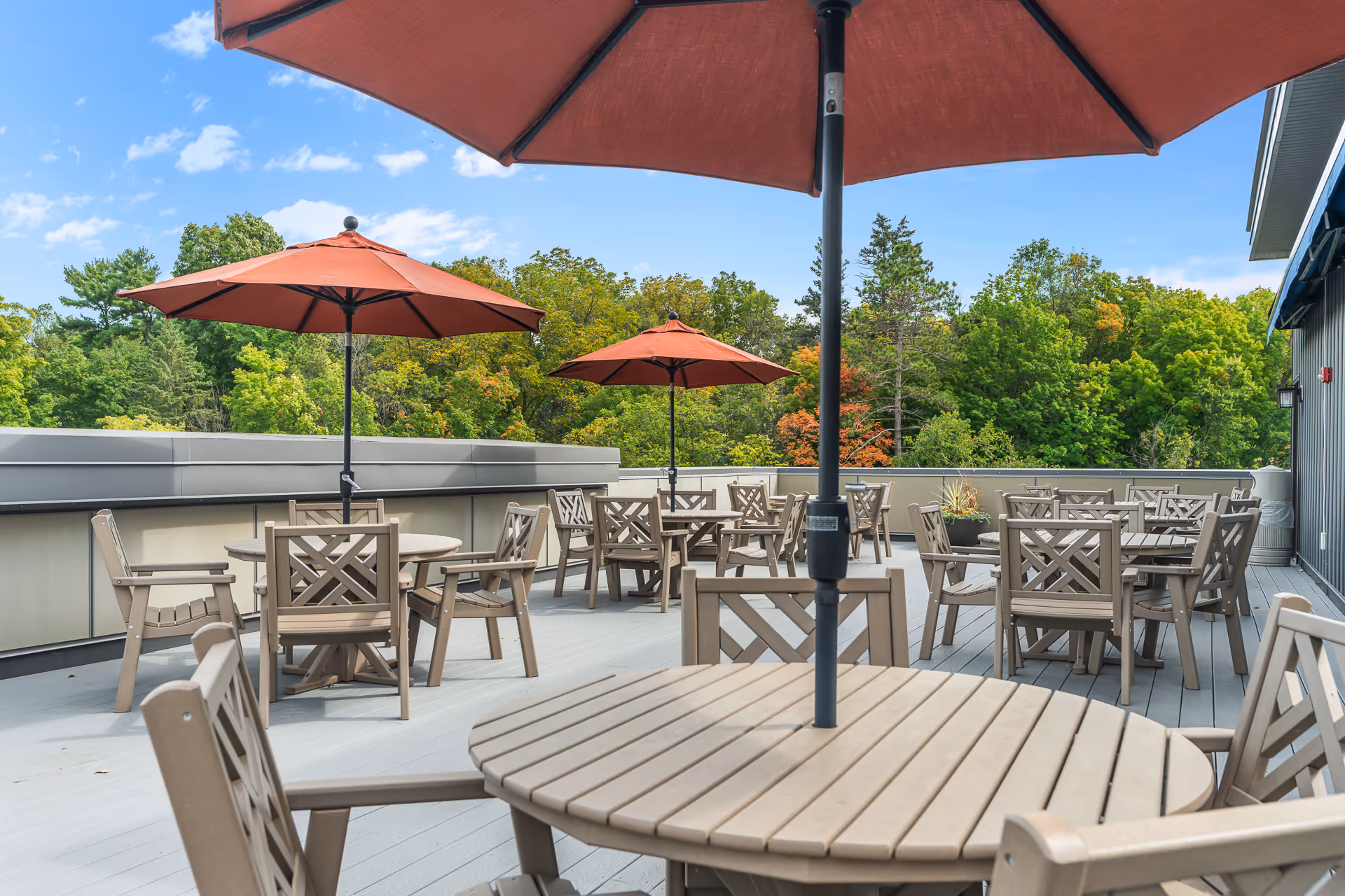 Outdoor patio area with multiple round tables and chairs made of light-colored wood. Each table has a large red umbrella providing shade. The patio overlooks a lush green area with trees under a clear blue sky.