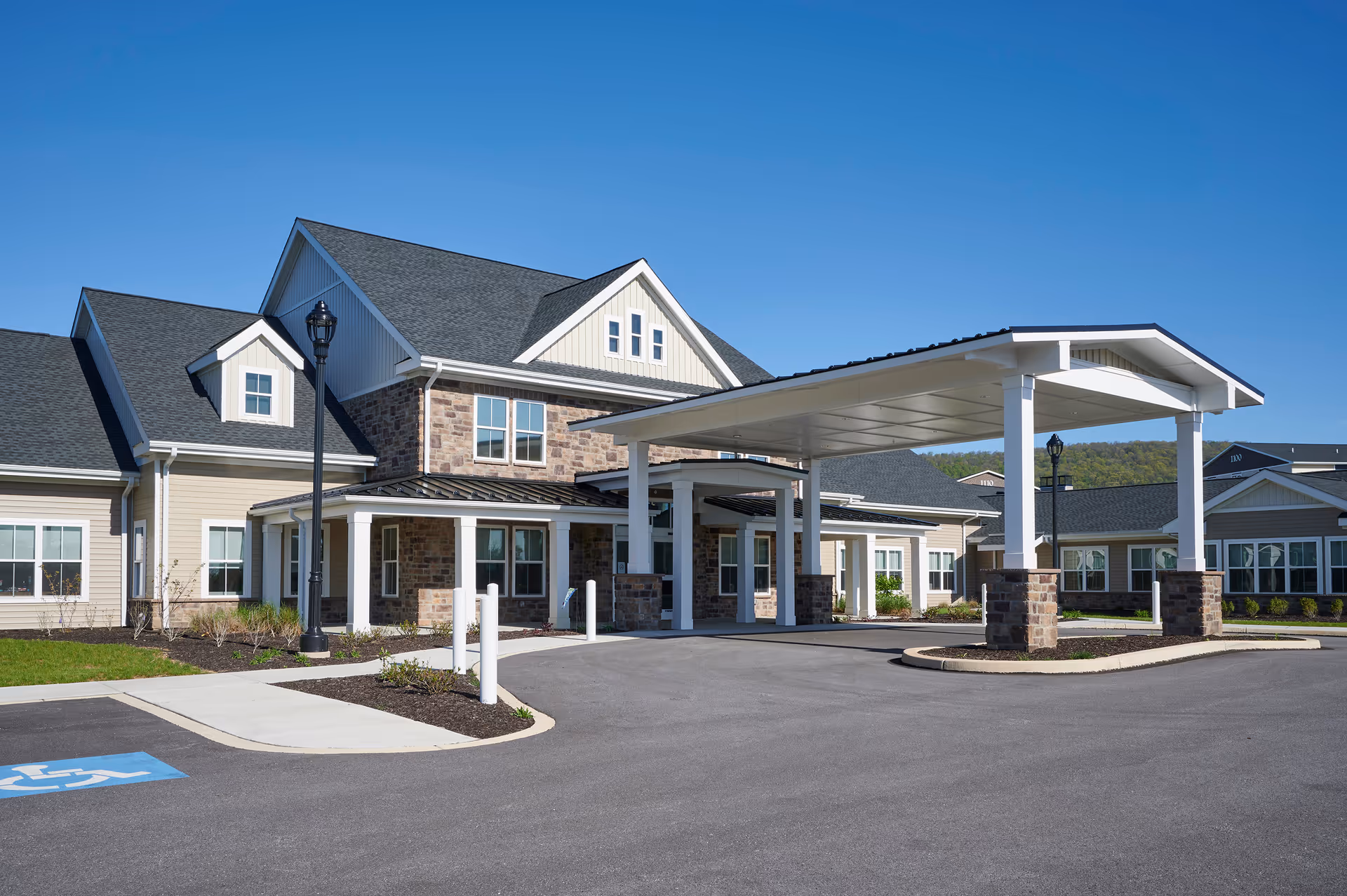 Front exterior of a two-story senior living facility with a covered drop-off porte-cochere and parking area under a clear blue sky.