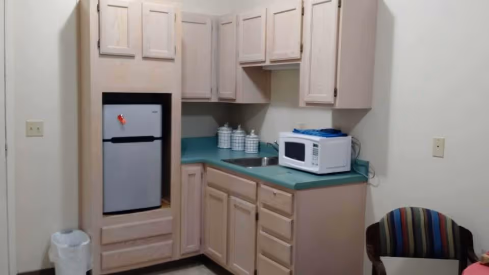 Small kitchenette area with light wood cabinets, a green countertop, a small refrigerator, a microwave, a sink, and three white canisters. A striped chair is partially visible on the right side and a white trash can is on the left side.
