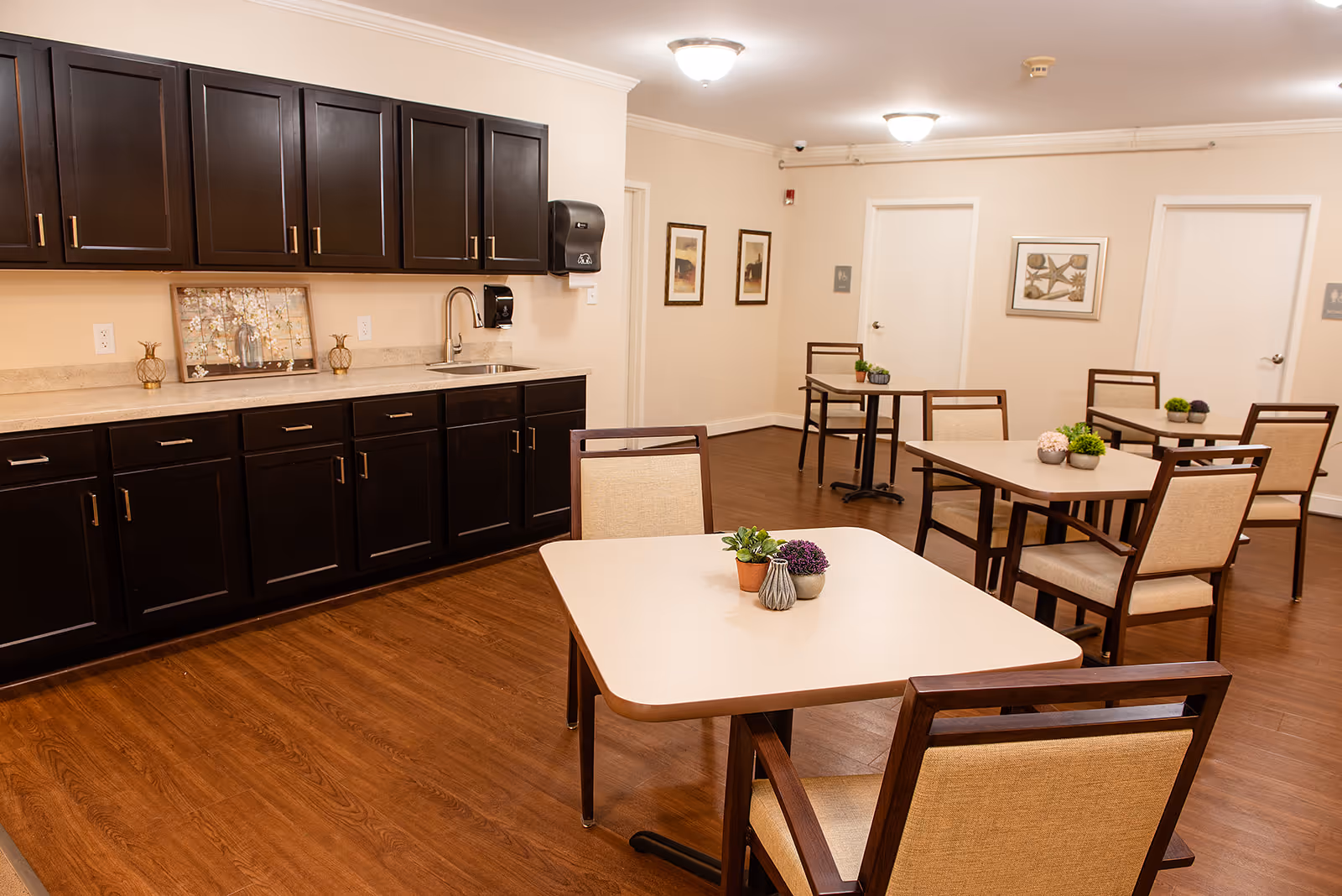A clean and well-lit dining area with several square tables and cushioned chairs. Each table has small decorative plants. Along one wall, there are dark wood cabinets with a countertop and a sink. The walls are light-colored with framed artwork and two doors visible in the background.