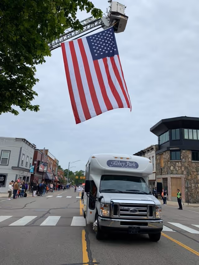 A white Abbey Park at Mill River shuttle bus is parked in the middle of a street during a parade or public event. A large American flag is hanging overhead from a fire truck ladder. People are gathered along the sidewalks on both sides of the street, and buildings line the street under a cloudy sky.