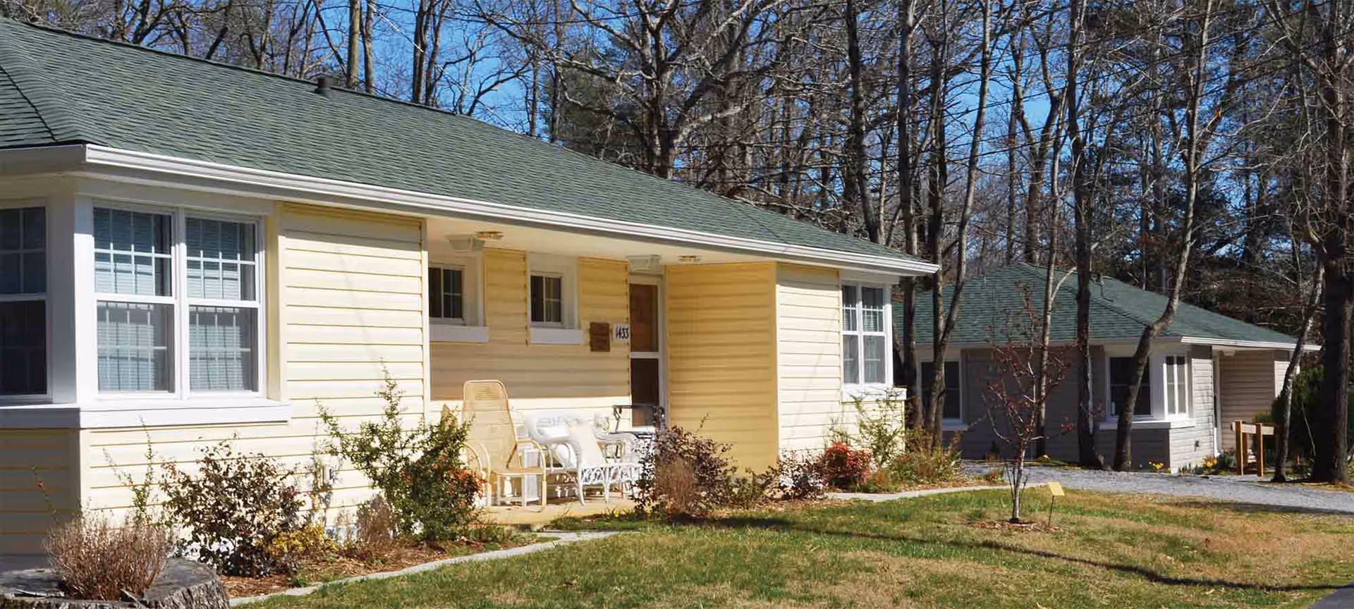 Single-story yellow cottage-style building with a small front porch, outdoor chairs, and a grassy yard surrounded by trees.