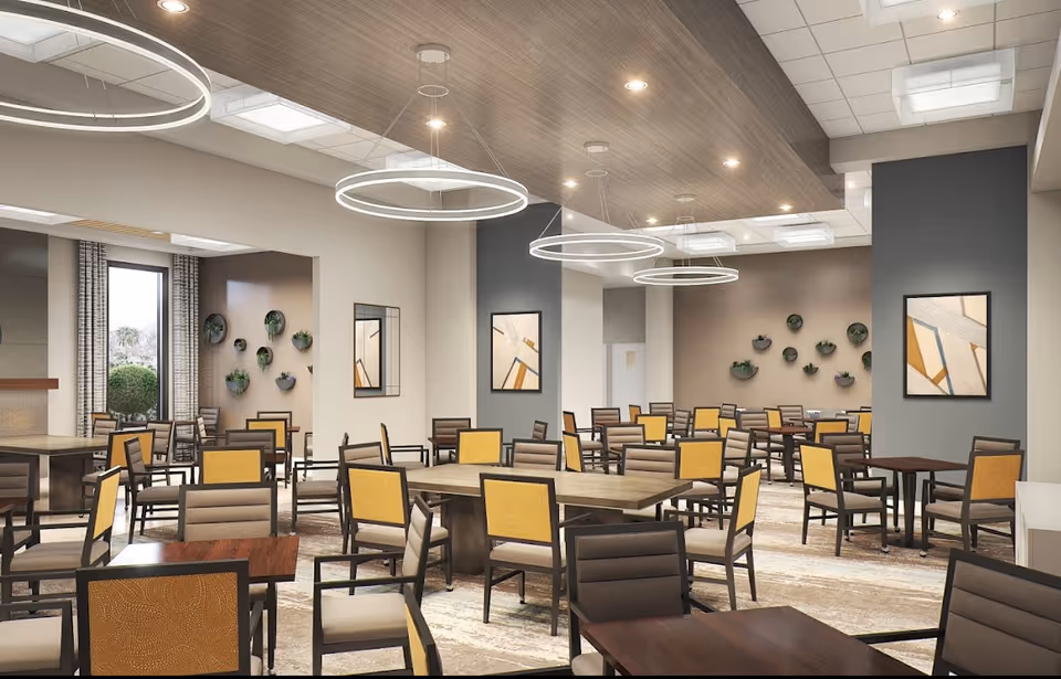 A spacious dining room with multiple tables and chairs arranged neatly. The chairs have cushioned seats and backs in beige and yellow tones. The room features modern circular pendant lights hanging from a wooden ceiling panel, abstract wall art, and small wall-mounted planters with greenery. Large windows allow natural light to enter the space.