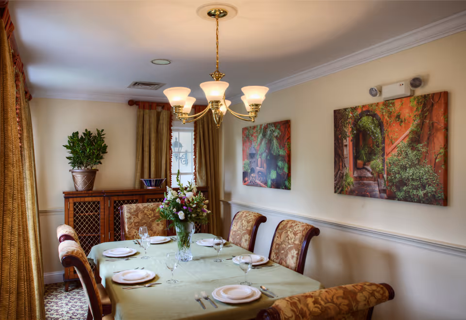 Formal dining room with a table set for six, a chandelier overhead, floral centerpiece, upholstered chairs, and framed artwork on the walls.