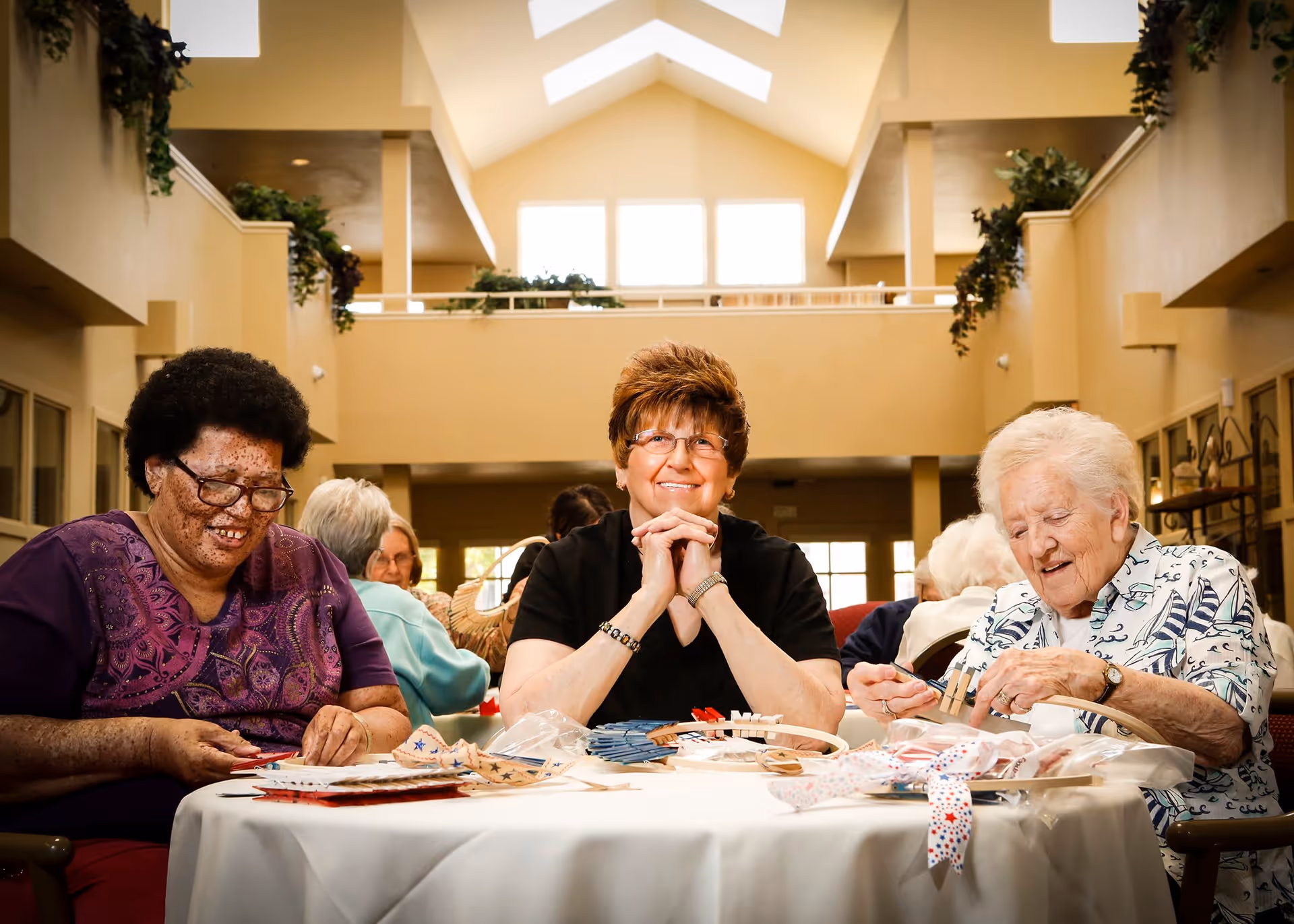 Three elderly women sitting around a round table engaged in a craft activity inside a spacious, well-lit room with high ceilings and large windows. The woman in the center is smiling and looking at the camera, while the other two women are focused on their crafts. The room has beige walls and greenery decorating the upper ledges.
